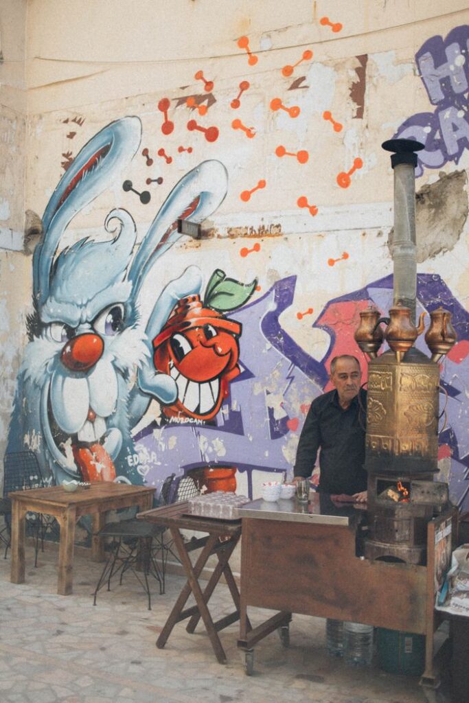 Man standing by a street-side tea stall against colorful graffiti art in an urban setting.